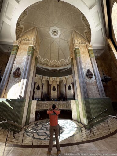 Wide-angle circular interior with patterned ceiling, marble columns, green tilework, gallery, floor medallion, and central visitor.