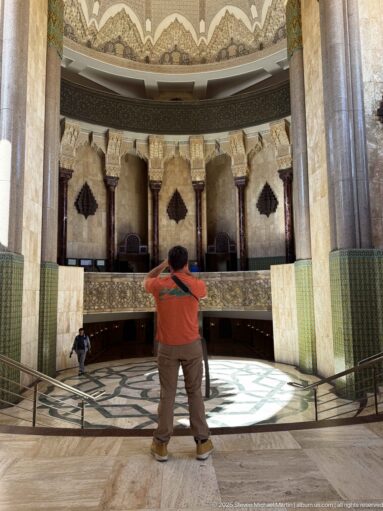 Visitor faces a circular mosque interior with marble columns, green tilework, carved capitals, gallery, and patterned floor.