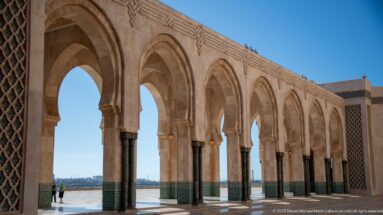 Repeated stone horseshoe arches and dark columns form an open colonnade beside the waterfront at the Hassan II Mosque.