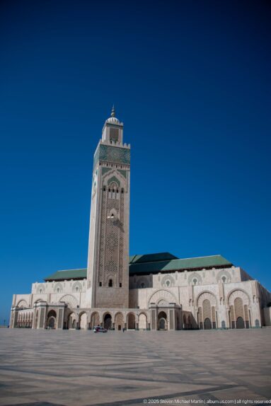 Low-angle mosque exterior across a broad plaza, with central minaret, arcades, and green roofline.