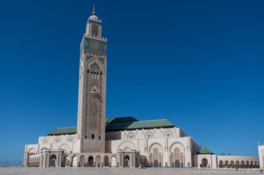 Wide Hassan II Mosque exterior with tall square minaret, green roofs, arcades, plaza, and small visitors.