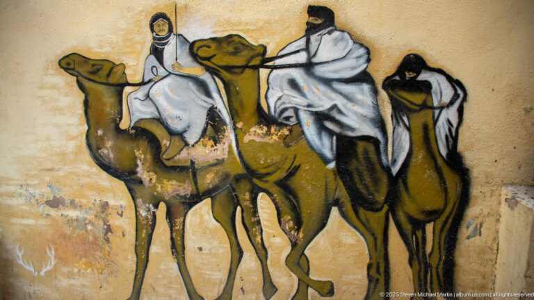 Mural of three camels carrying white-robed riders across an ochre wall.
