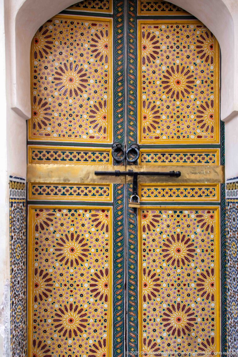 Brightly painted double wooden door with floral starburst patterns, dark metal hardware, and narrow zellij side panels.