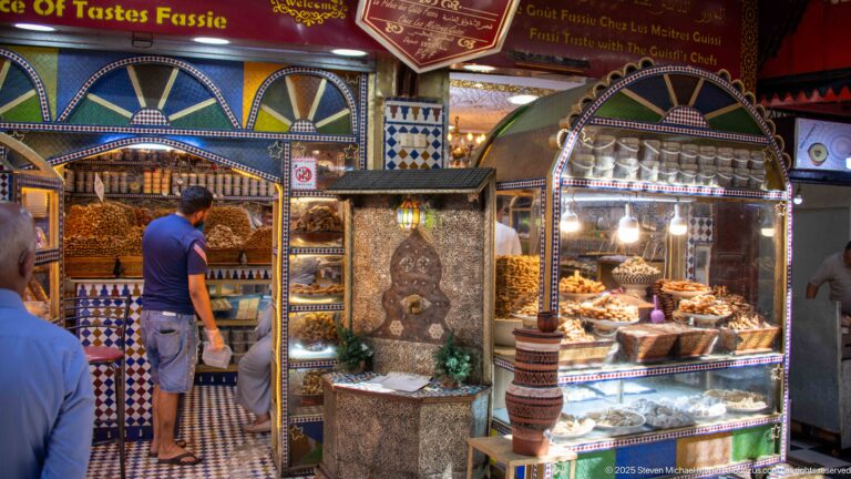 Customer at an ornate sweets shop with trays of pastries, glass display cases, colorful arches, and patterned tilework.