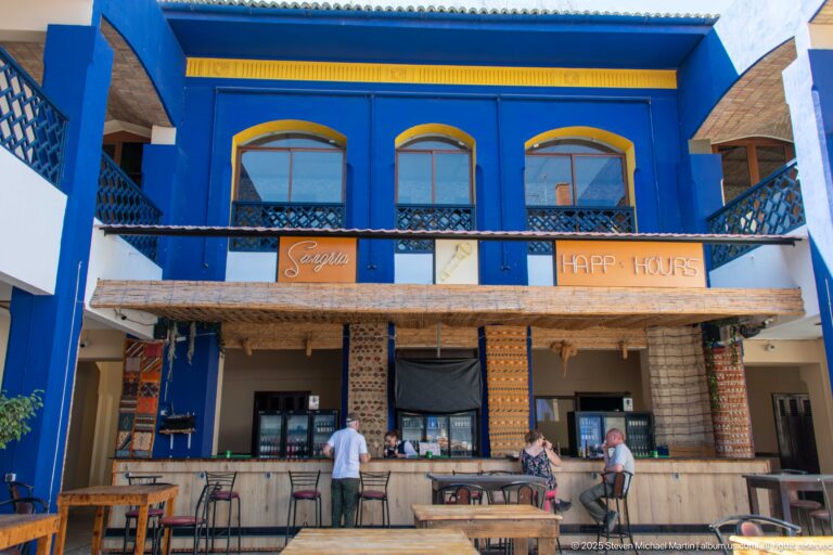 Blue two-story café façade with yellow trim, balcony railings, wooden counter, and patrons below.