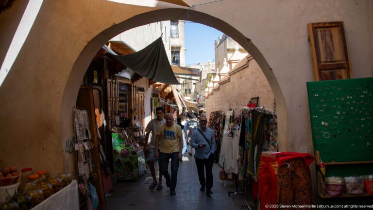 Two pedestrians walk through a broad arch into a narrow market lane lined with textiles and shop goods.