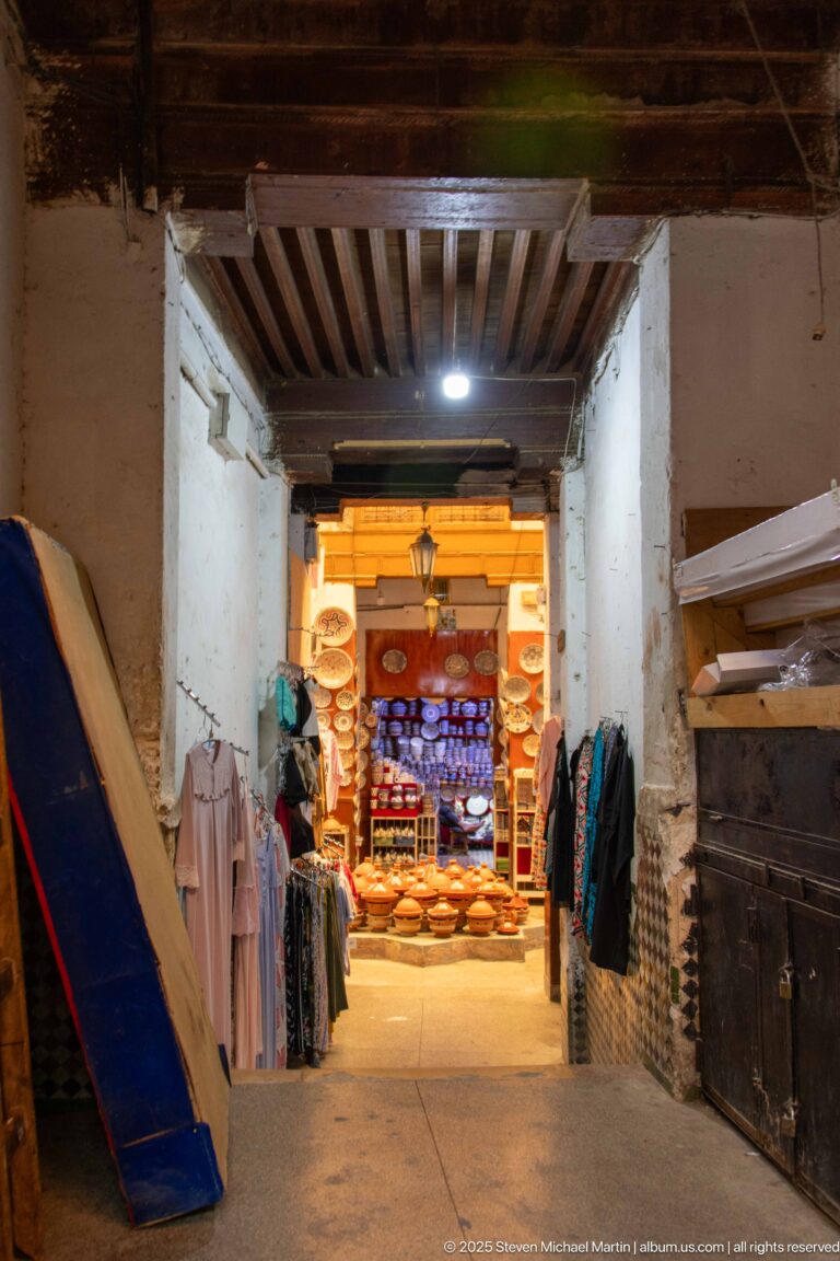 Dim timber-roofed market passage leading to a bright shop, with clothing and goods lining the walls.