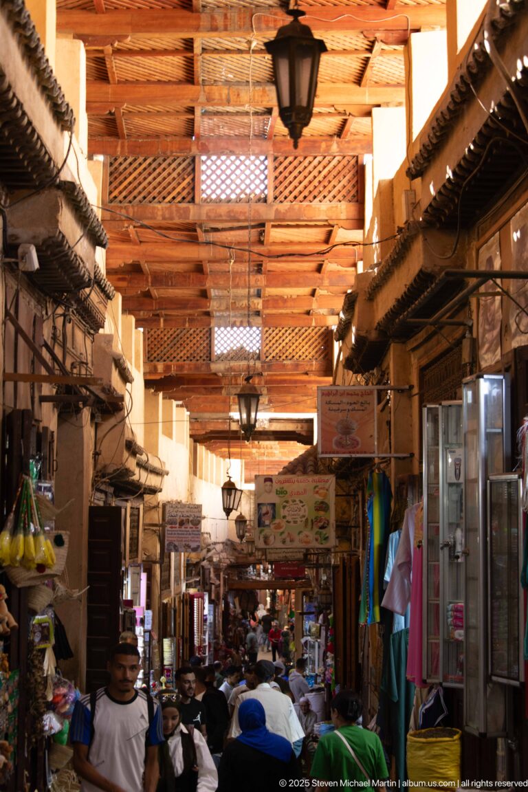 Busy covered market street beneath timber beams, with hanging lamps, tightly packed shops, awnings, and pedestrians.