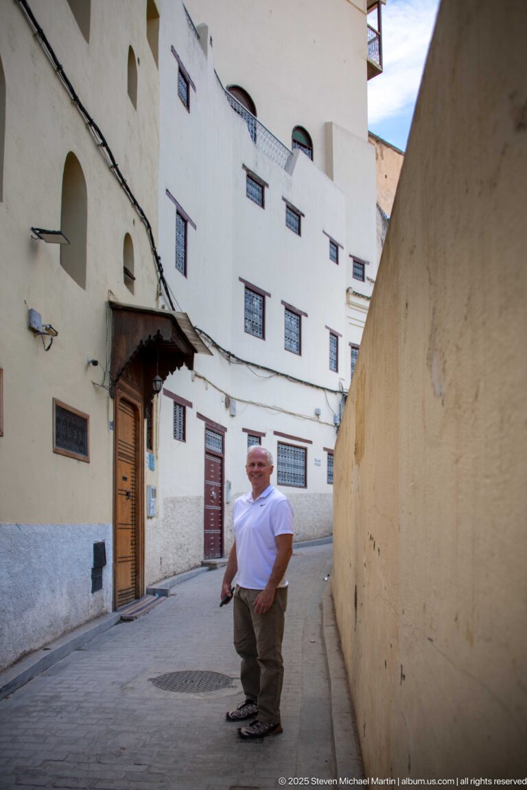 Visitor standing in a narrow medina lane beside pale façades, barred windows, wooden doors, and projecting awnings.