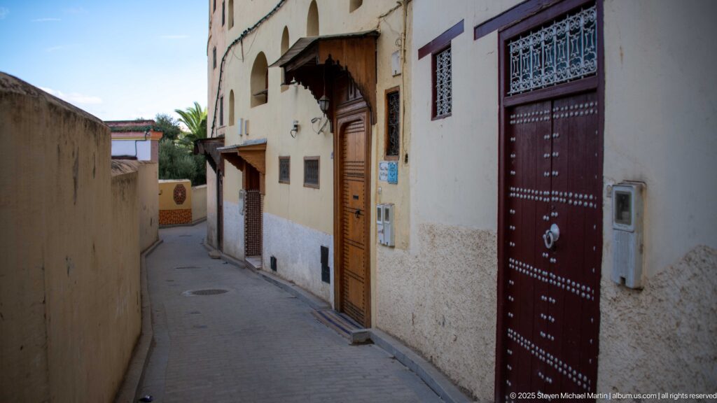 Narrow residential lane lined with heavy wooden doors, barred windows, awnings, and pale plaster walls.