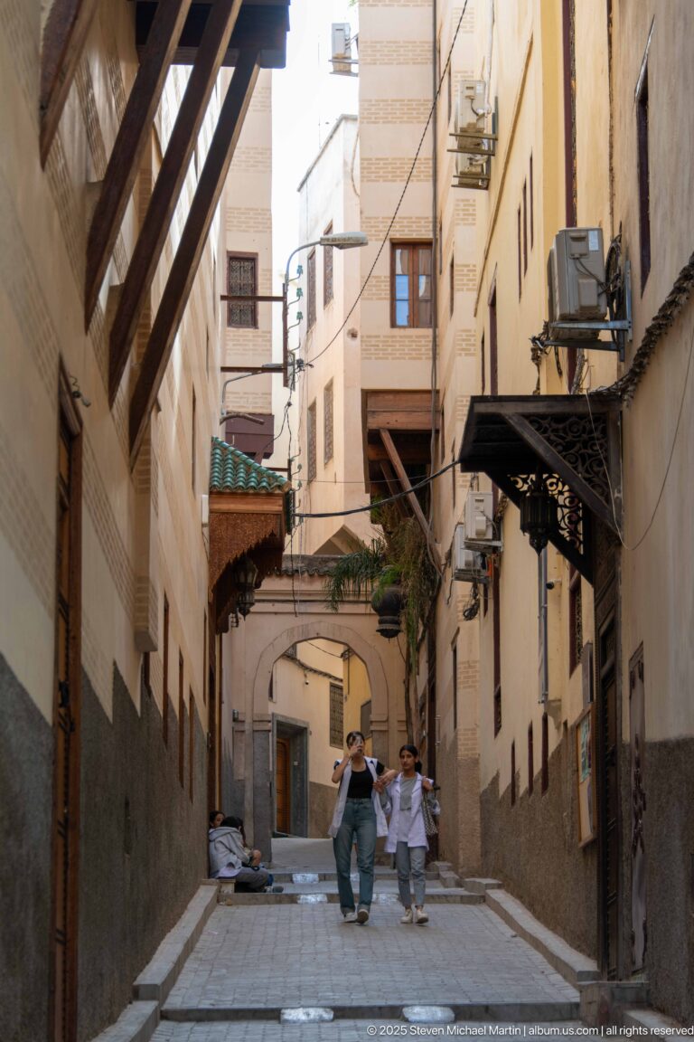 Pedestrians walking along a narrow medina street beneath projecting structures and arched building connections.
