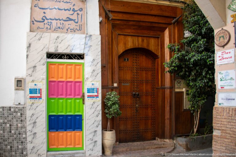 Bright multicolored small door beside a large studded wooden entrance framed by tile, signs, and greenery.