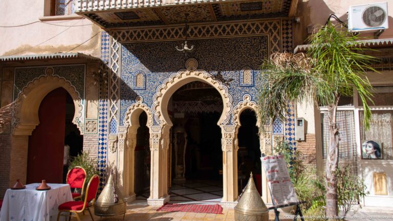 Ornate multi-arched entrance with zellij, carved plaster, dark patterned walls, metal vessels, and a red chair.