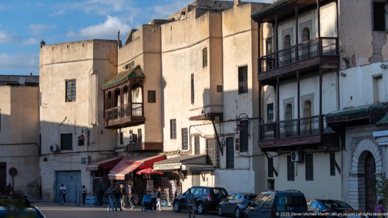 Street with projecting wooden balconies, shops, pedestrians, and parked cars beside tall weathered medina buildings.
