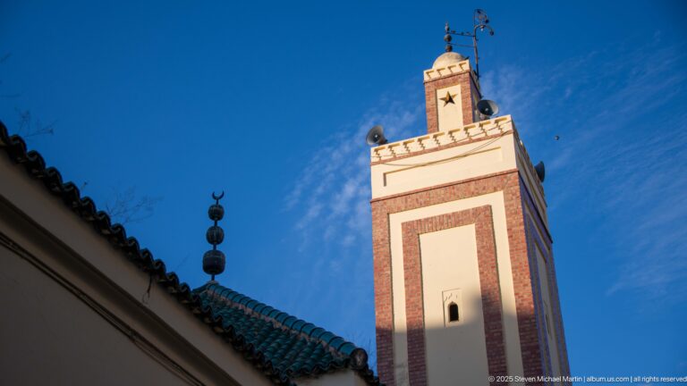 Square mosque minaret with reddish vertical panels rising above a green-tiled roof against blue sky.
