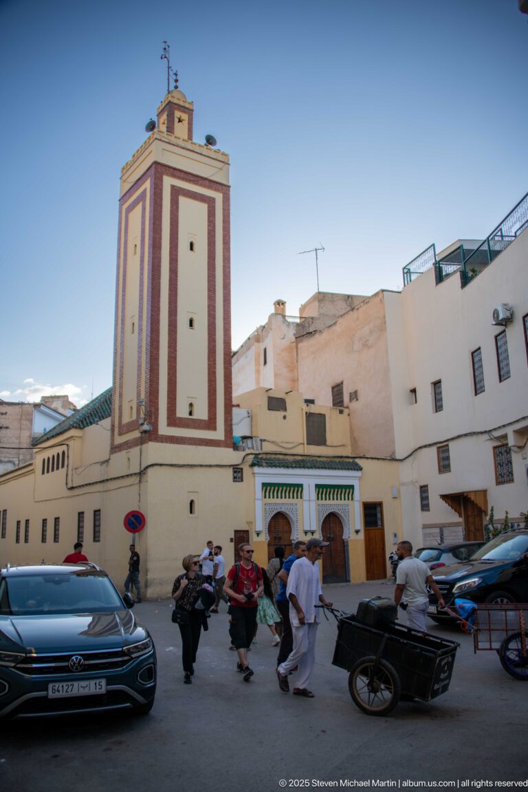 Square mosque minaret above a busy street with pedestrians, vehicles, motorbikes, and a horse-drawn cart.