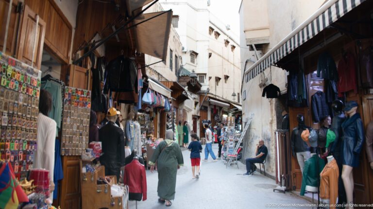 Busy narrow souk lane lined with hanging clothing, souvenirs, shop displays, awnings, and pedestrians.