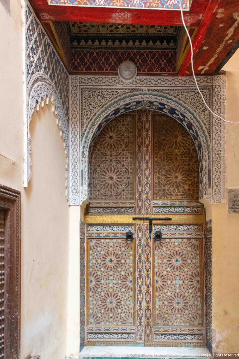 Polychrome double wooden door covered with geometric star patterns beneath an ornate arched surround.