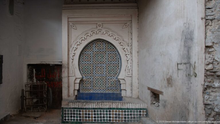 Small arched wall fountain with blue-and-white mosaic tile set in a worn plaster passage beside stacked metal furniture.