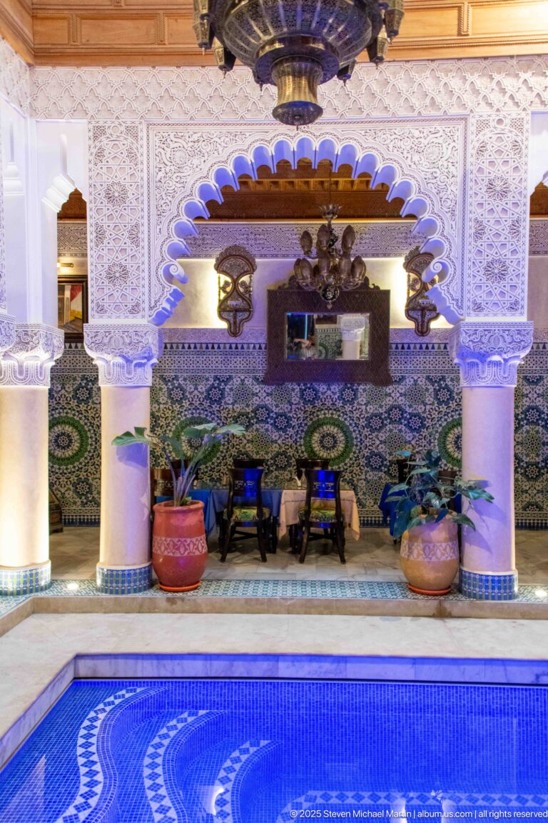 Blue-lit pool facing an ornate lounge with scalloped arch, columns, zellij, carved plaster, lanterns, and large planters.