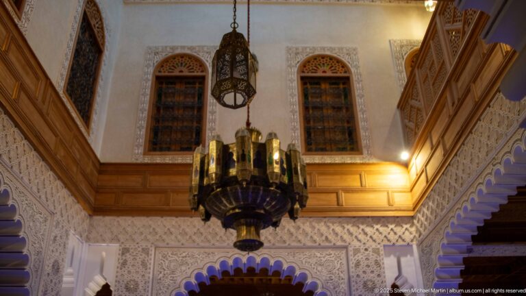 Hanging metal lanterns beneath tall screened windows, carved plaster, wood trim, and patterned arches in a Fes riad interior.