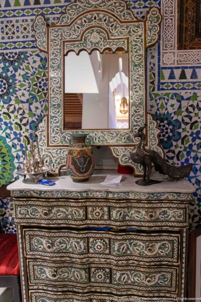 Inlaid chest of drawers and framed mirror against a wall covered with blue, green, and white geometric zellij.