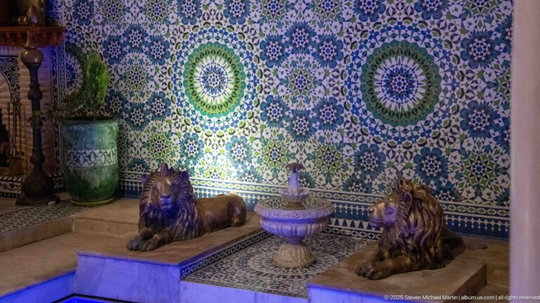 Two reclining lion sculptures beside a small pedestal fountain against a wall of intricate blue, green, and white mosaic.