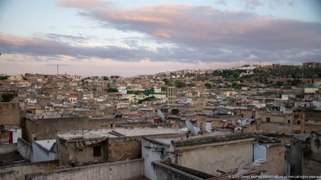 Elevated view over dense flat-roofed buildings of Fes Medina beneath a cloud-streaked evening sky.