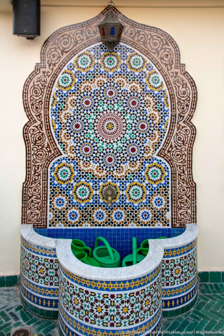Horseshoe-arched wall fountain covered in multicolored geometric mosaic, with green watering cans resting in its tiled basin.