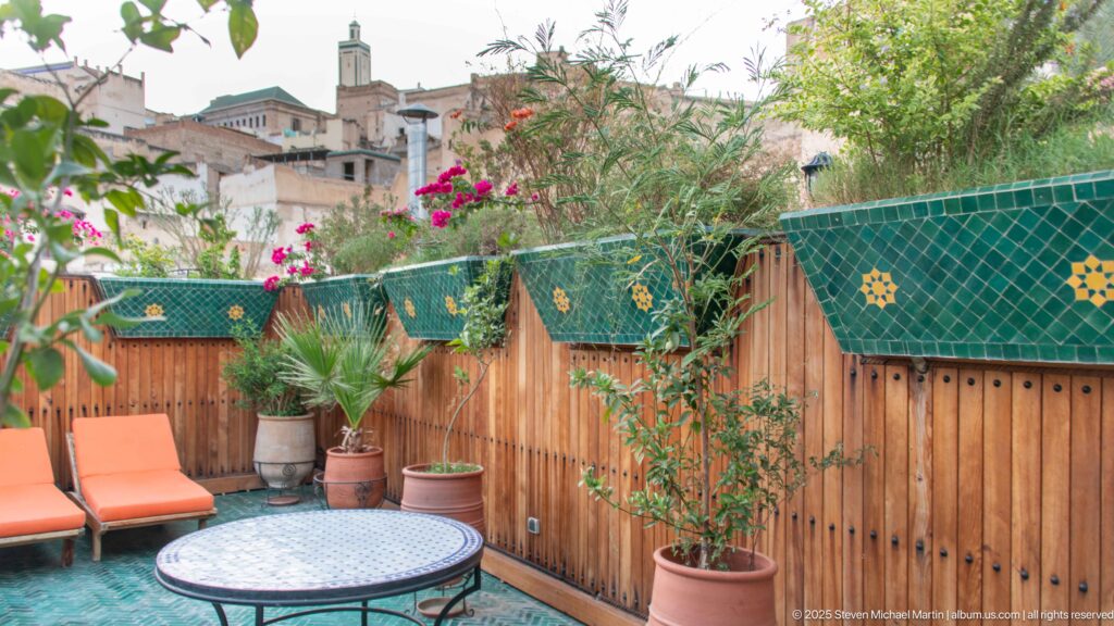 Potted plants, orange chairs, and a round mosaic table on a terrace overlooking masonry buildings in Fes Medina.
