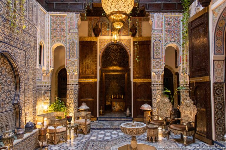 Ornate riad courtyard with central fountain, patterned floor, carved arches, tilework, dark wood, lamps, and seating.