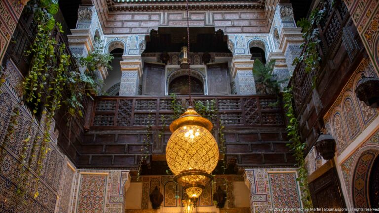 Large glowing lantern below a dark carved-wood gallery, with decorated arches and hanging plants in a Fes courtyard.