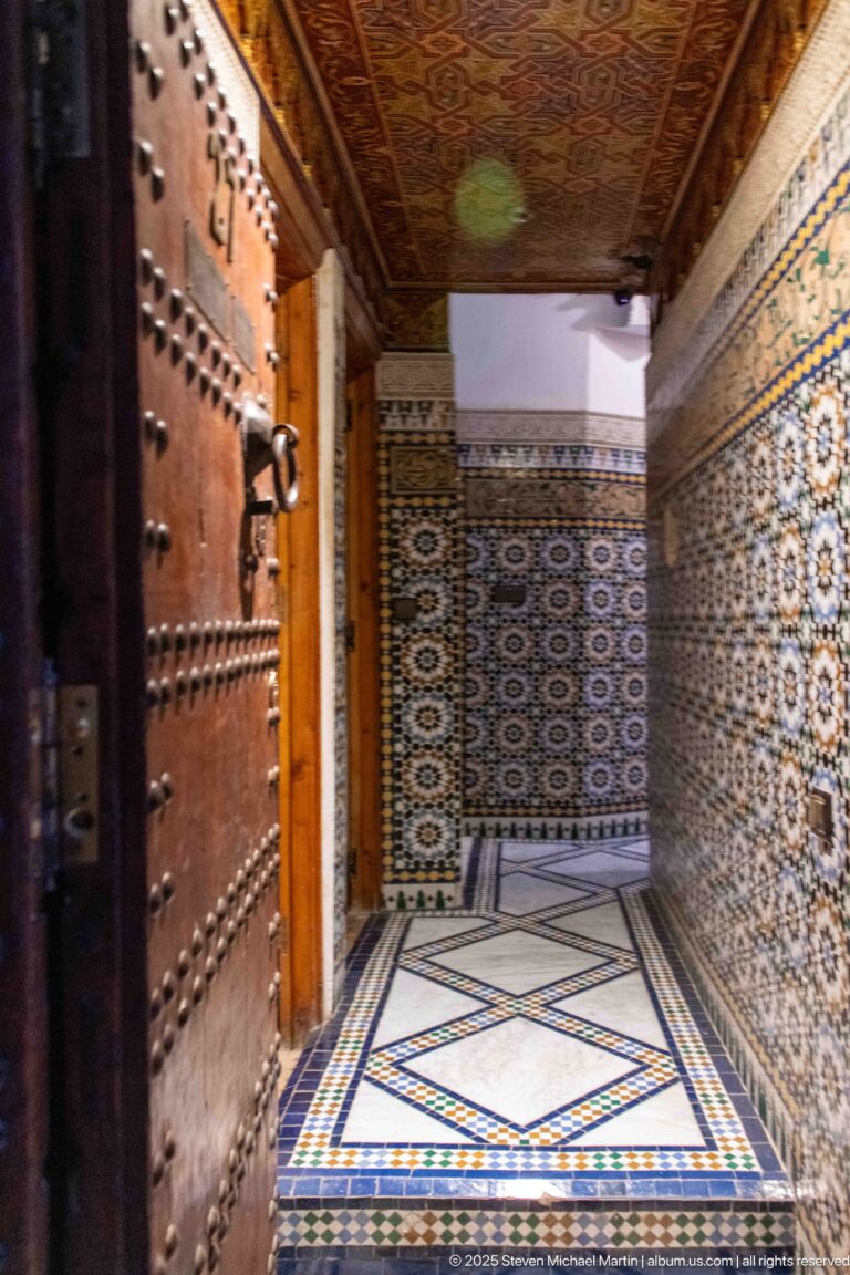 Studded wooden door opening into a narrow tiled passage with geometric floor and decorated wooden ceiling.