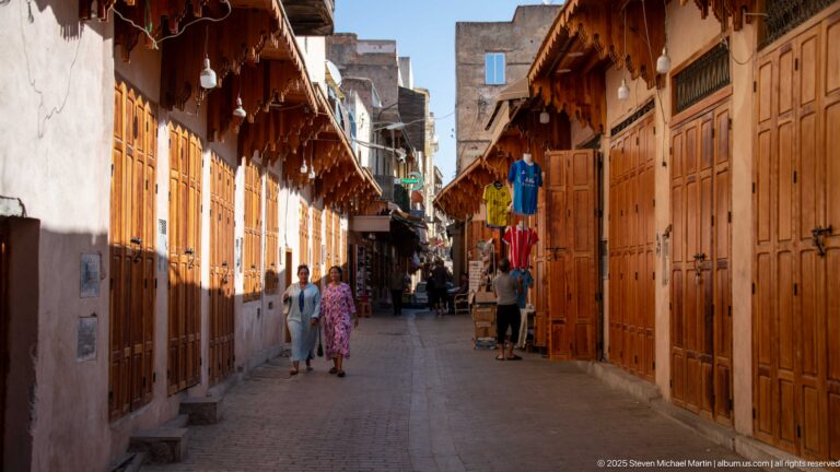 Pedestrians walking along a narrow Fes Medina lane lined with tall wooden shop doors and projecting awnings.