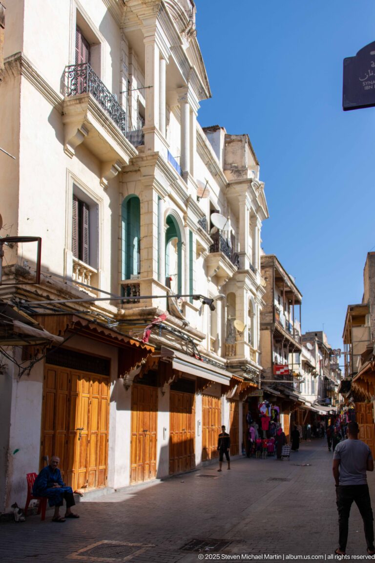 Sunlit commercial street in Fes with wooden shopfronts, white upper façades, balconies, green shutters, and pedestrians.
