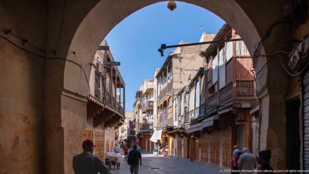 View through a shaded masonry arch toward a sunlit Fes street with pedestrians, balconies, awnings, and shopfronts.