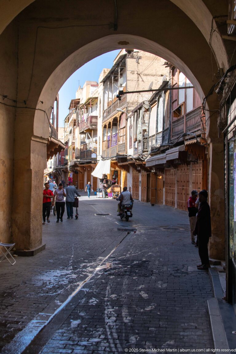 Large shaded arch framing an open paved market area with pedestrians, balconies, awnings, and wooden shopfronts.