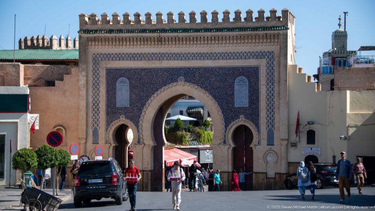 Blue-tiled Bab Bou Jeloud gate with central horseshoe arch, two smaller openings, crenellated top, pedestrians, and vehicles.