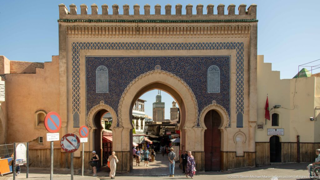 Frontal view of blue-tiled Bab Bou Jeloud with pedestrians passing through and a minaret visible beyond the central arch.