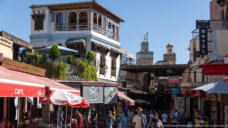 Busy Fes Medina street with café awnings, pedestrians, a projecting windowed upper room, and minarets in the distance.