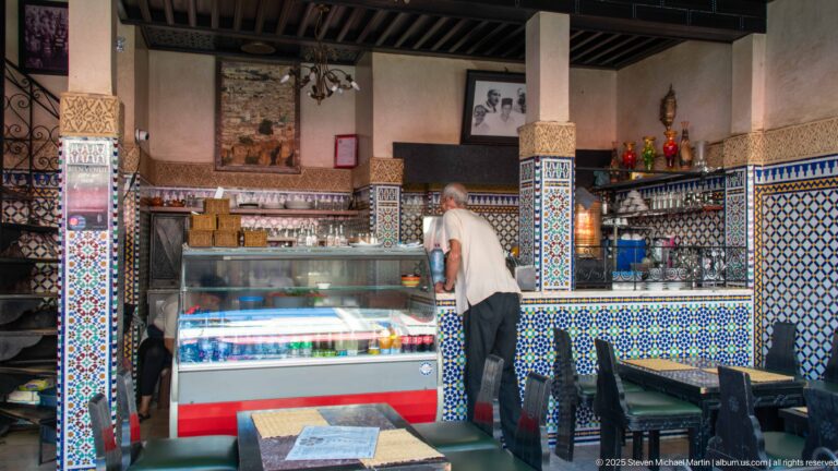 Man at a refrigerated display counter in a small café decorated with blue-and-white geometric tiles and patterned columns.