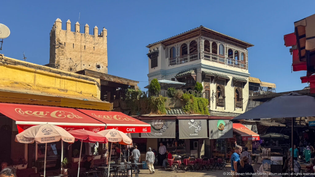 Cafés and pedestrians beneath a crenellated tower and projecting windowed building in a sunny Fes Medina streetscape.