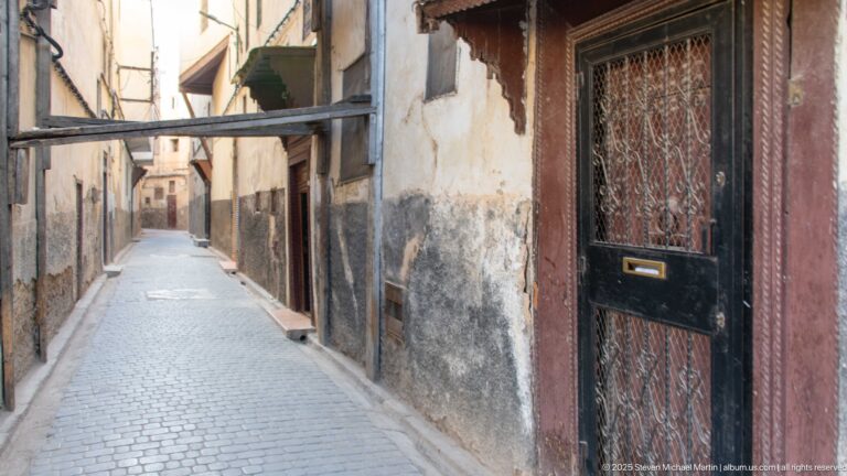 Long narrow medina lane with worn plaster walls, doorways, cobbled paving, and timber braces overhead.