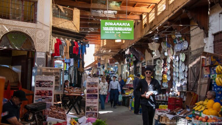 Shoppers walking through a narrow covered souq lined with fruit, cookware, clothing, signs, and small market stalls.