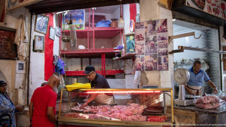 Men working at a butcher stall with raw meat in a glass counter, hanging scale, and preparation area.