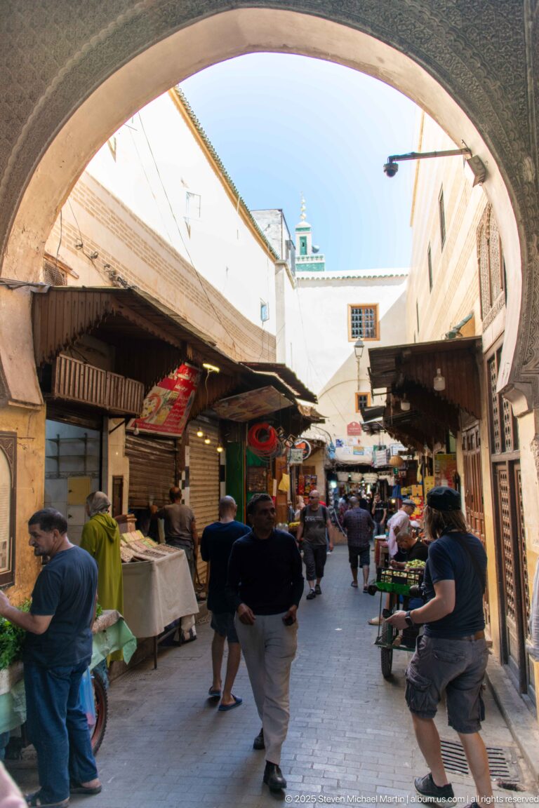 Pedestrians passing beneath a broad arch into a Fes market lane lined with produce tables and covered stalls.