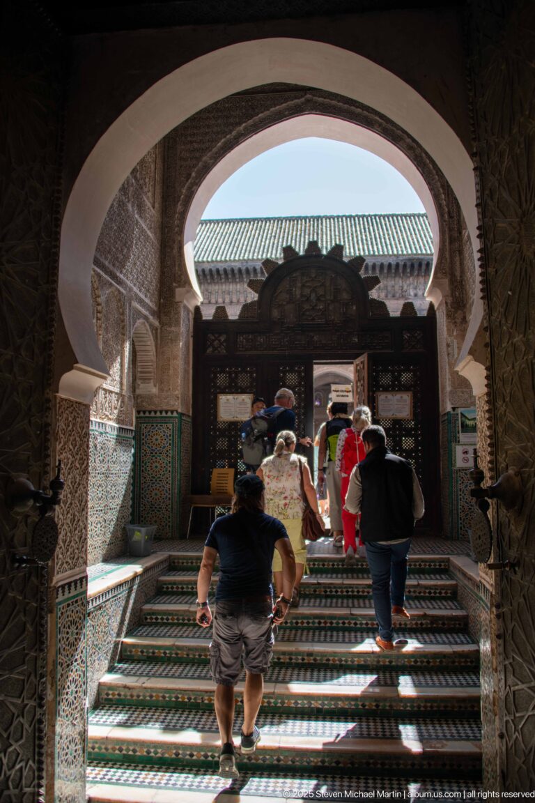 Visitors climbing patterned tiled steps beneath a horseshoe arch toward an elaborate dark wooden screen in a decorated interior.