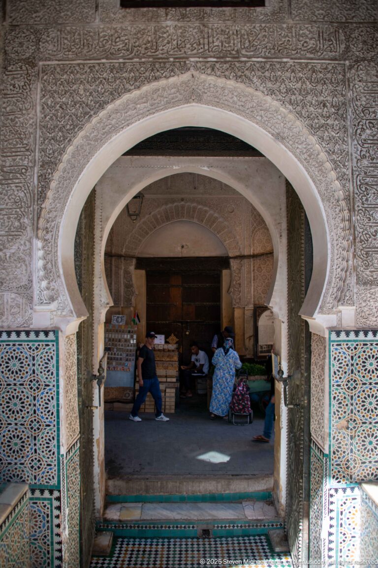 Nested horseshoe arches with carved plaster and blue-green geometric tilework opening into a shaded historic interior.