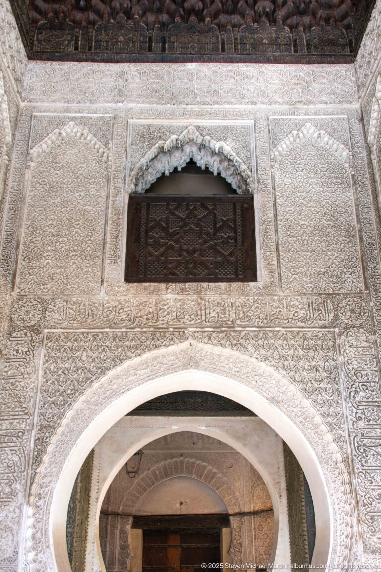 Carved plaster wall above a horseshoe arch with dark wooden lattice window, scalloped hood, and blue-green tilework below.