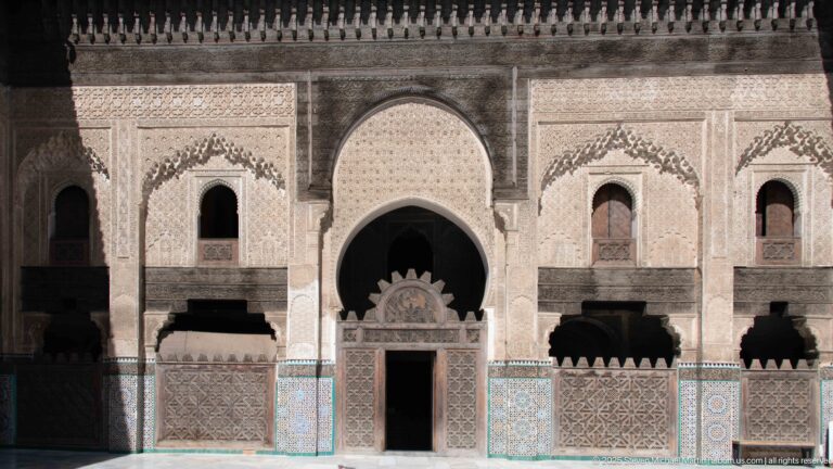 Symmetrical decorated courtyard wall with central arched doorway, smaller windows, carved wood screens, pale plasterwork, and blue-green zellij.